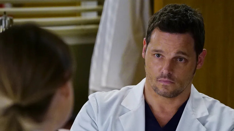 Justin Chambers as Dr. Alex Karev looking worried with a white lab coat and black scrubs on while talking to a female character with her hair in a ponytail who we only see from behind in Grey's Anatomy