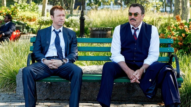 Donnie Wahlberg as detective Danny Reagan, wearing a blue suit and white collared shirt, sitting on a green park bench next to Commissioner Frank Reagan, wearing a dark navy vest and pants with a jacket on his knee and a blue and red striped tie, in Blue Bloods