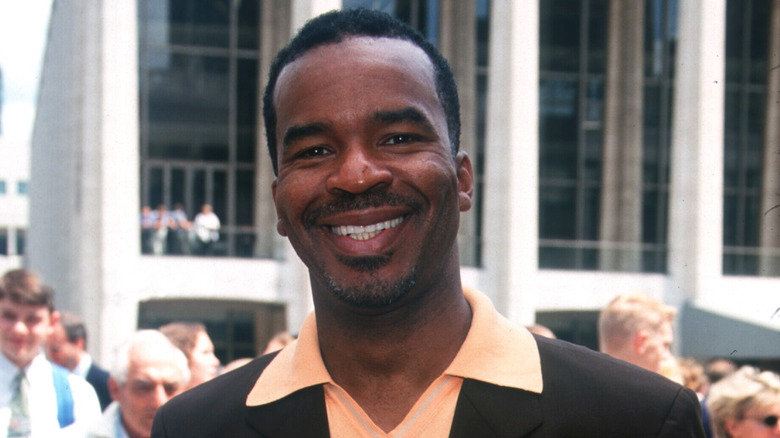 David Alan Grier, wearing a brown jacket and peach colored collared shirt, smiling outside at a promotional event