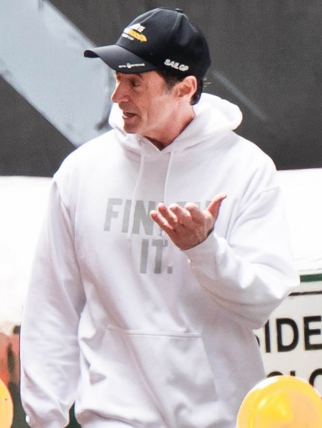 Hugh Jackman in a white hoodie with his hand raised