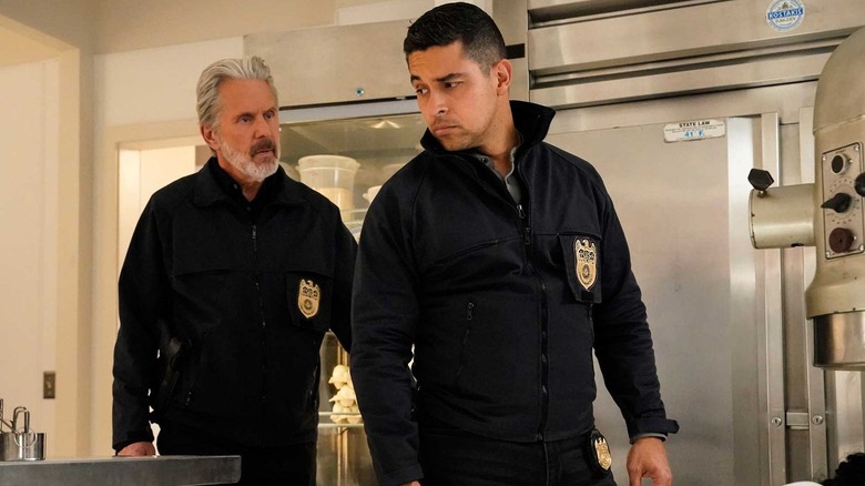 Gary Cole as special agent Alden Parker walking behind Wilmer Valderrama as special agent Nicholas Torres, who looks back over his shoulder at Parker, both wearing their NCIS black pullovers with badges over the left breast pocket, in an industrial kitchen in 