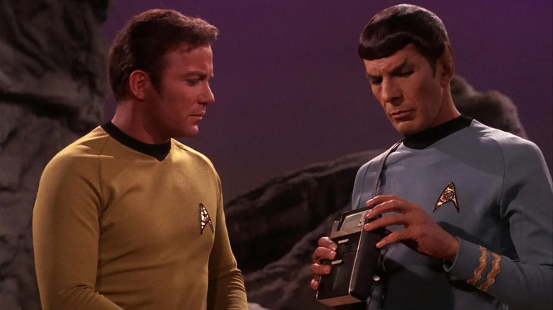 Captain James T. Kirk (William Shatner) stands next to Mr. Spock (Leonard Nimoy), who is holding a Tricorder in 