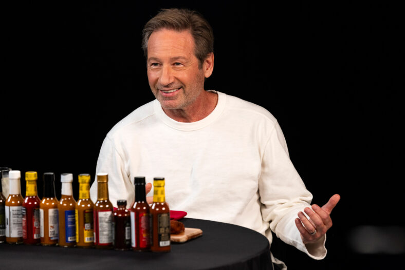 David Duchovny appears on Hot Ones, sitting behind a lineup of hot sauces while wearing a white sweater.