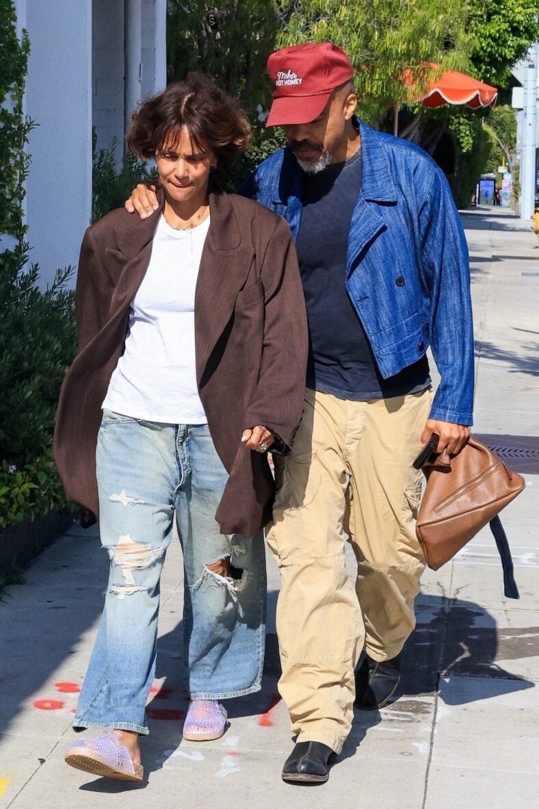 Halle Berry and fiancé Van Hunt walk arm-in-arm on a sidewalk, with Van wearing a bright blue jacket and red cap while Halle wears an oversized brown blazer and jeans