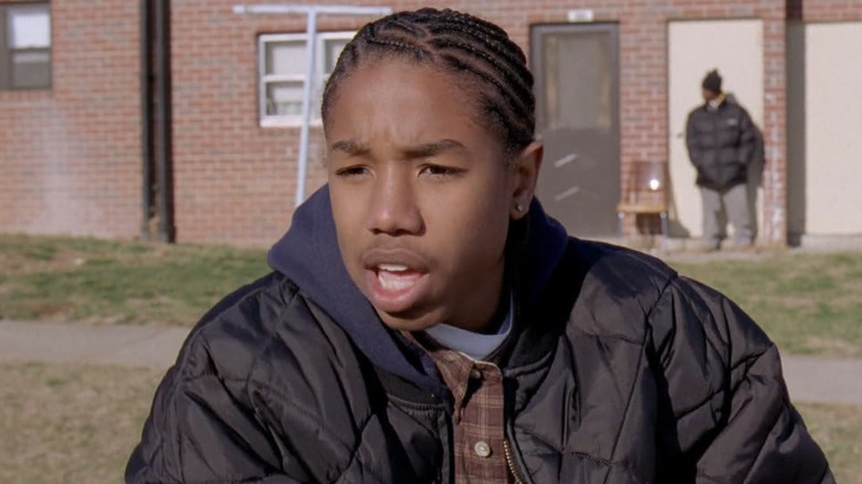 Michael B. Jordan wearing a quilted black jacket with a blue hood and a plaid shirt underneath as Wallace in 