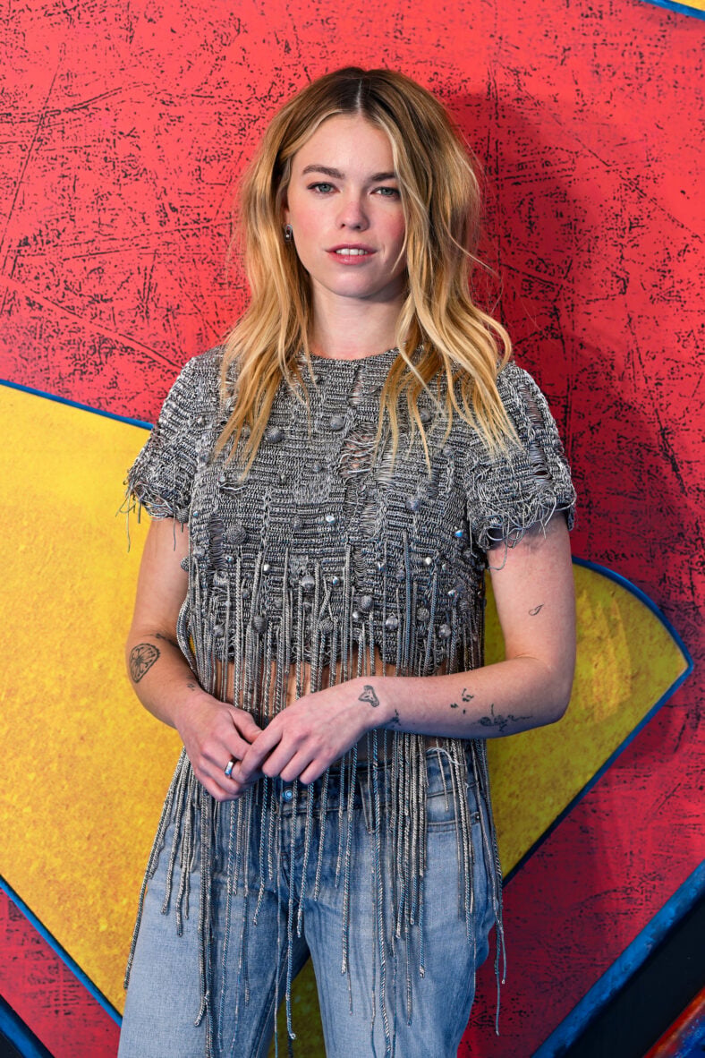 Milly Alcock poses against a red, yellow, and blue Supergirl backdrop while wearing a textured silver-gray fringe top, jeans, and showing several visible arm tattoos.
