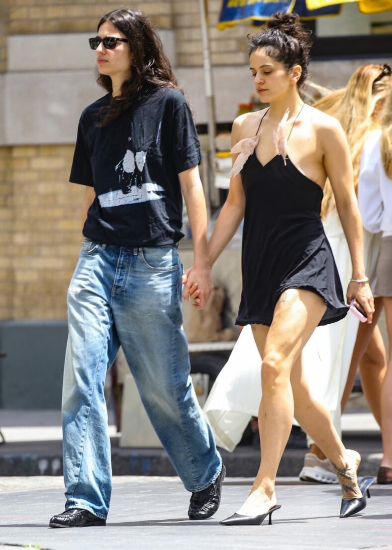Rosalia and Loli Bahia hold hands while walking together. Rosalia wears a black mini dress with her hair in a bun. Loli wears a black graphic tee with sunglasses.