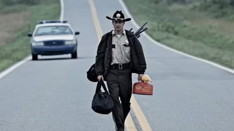 Rick Grimes walking down an empty road with supplies in hand and on his back, leaving his police car behind in the background