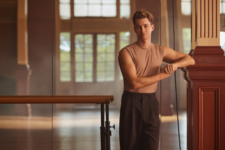 Dancing with the Stars: The Next Pro contestant photo featuring AJ Pritchard. He's in a tan sleeveless shirt, looking at the camera with a straight face, and leaning against a wood pillar. There's a mirror behind him and a dance bar to the left
