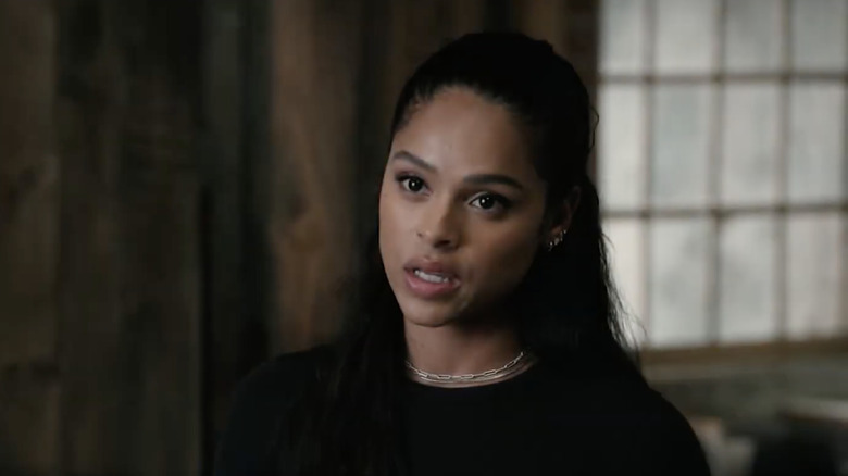 Ash Santos as Andrea Cruz. She has long black hair, several silver piercings in her ear, and she's wearing a silver necklace. She's wearing a black top and is stood in a wooden room with large window behind her.