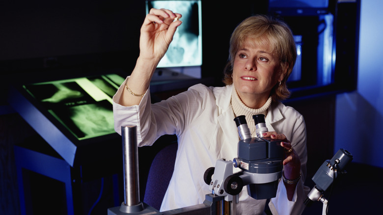 Dr. Kathy Reichs in a lab coat holding a tooth up in one hand and holding a microscope with the other.
