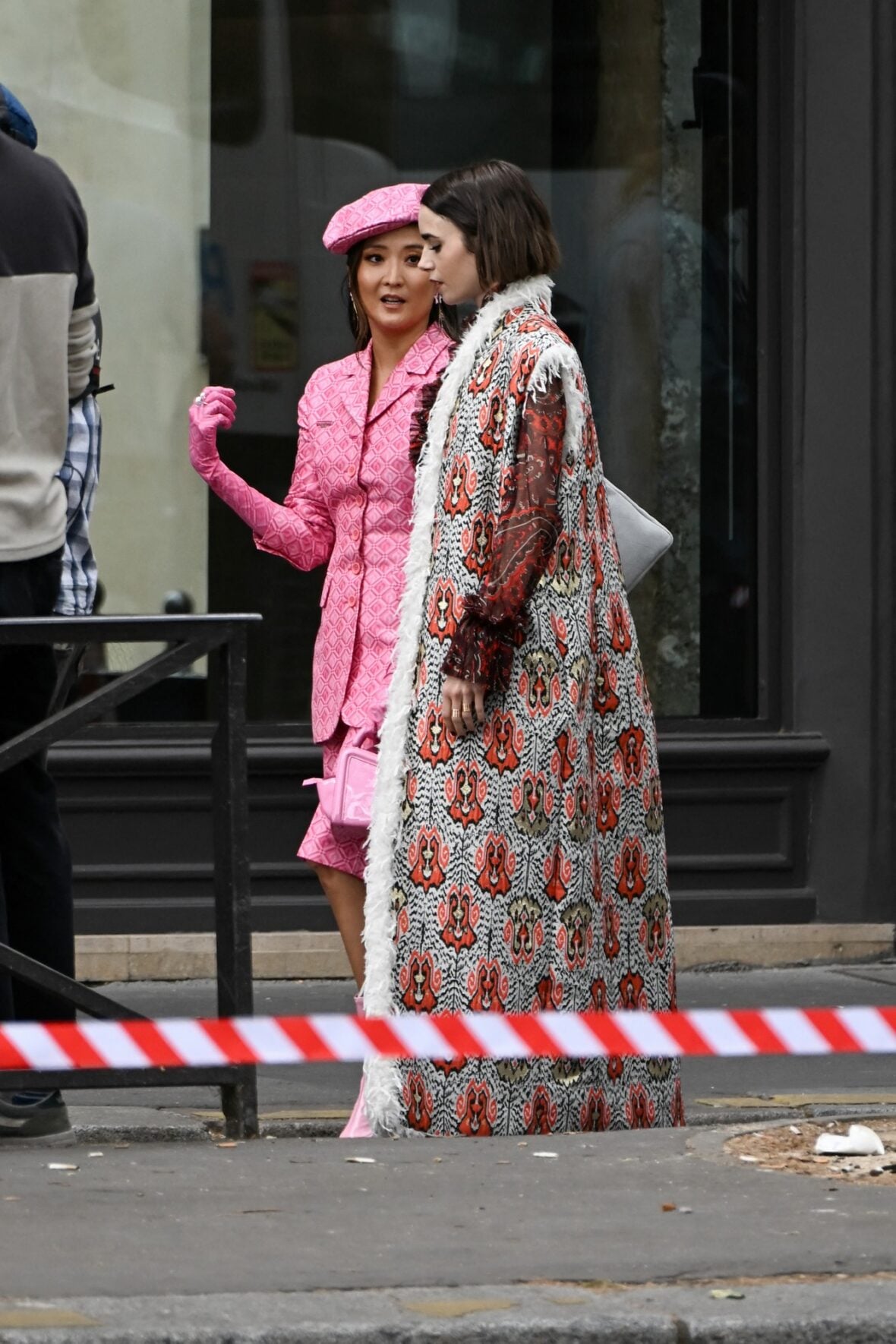 Ashley Park talks with Lily Collins on the set of Emily in Paris, with Ashley in a pink beret and matching suit and Lily in a long patterned coat.