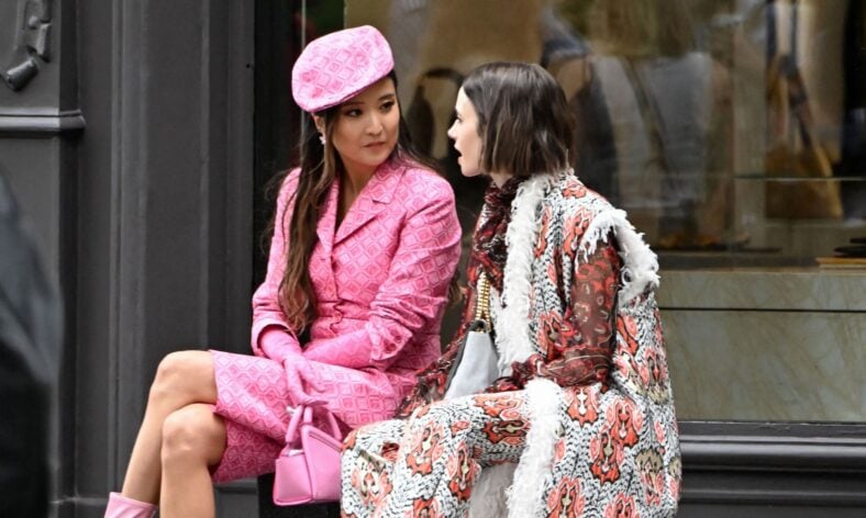 Lily Collins and Ashley Park sit together while filming Emily in Paris, with Ashley in a full pink look and Lily wearing a bold red, black, and white coat.