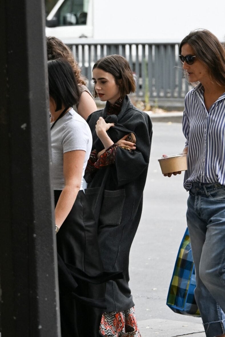 Lily Collins walks on the Paris set of Emily in Paris wearing a gray coat over her patterned outfit while holding her phone.