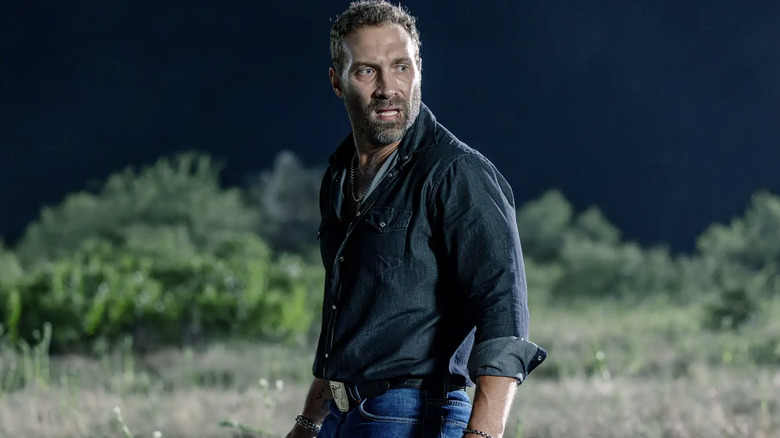 Jai Courtney as Will-Rob Jackson, he has short dark hair and a short beard. He's wearing a dark blue shirt and light blue denim jeans.