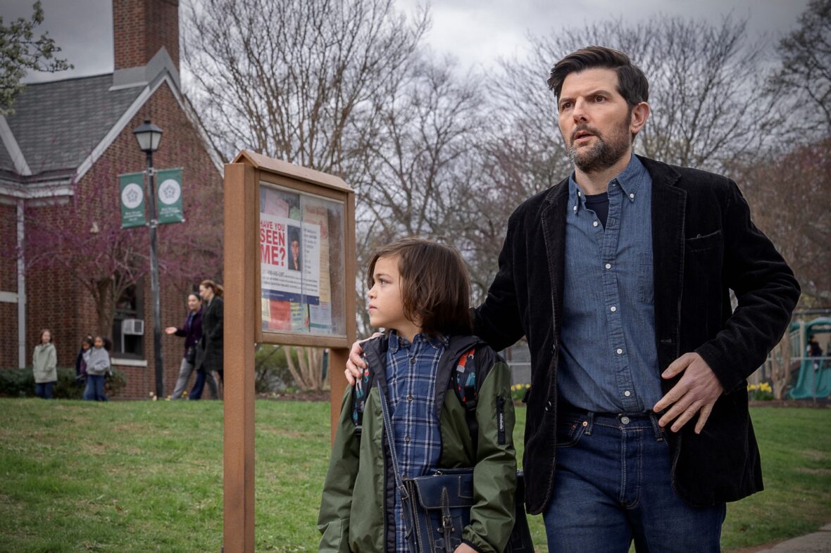 Adam Scott with his arm around the actor playing his son, Acston Luca Porto, as they stand outside a school. A missing flyer for another boy is seen on a bulletin board behind them. Still from