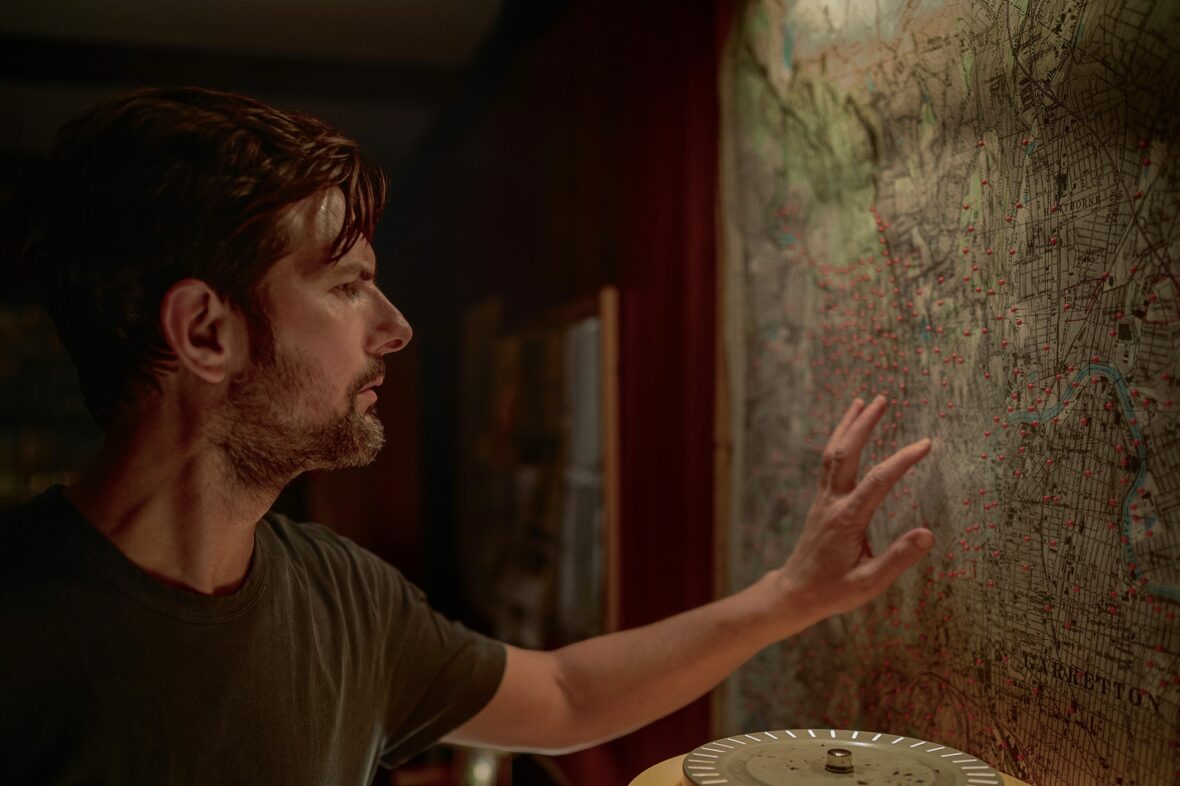 Adam Scott puts his hand up to a map covered in pins in a still from