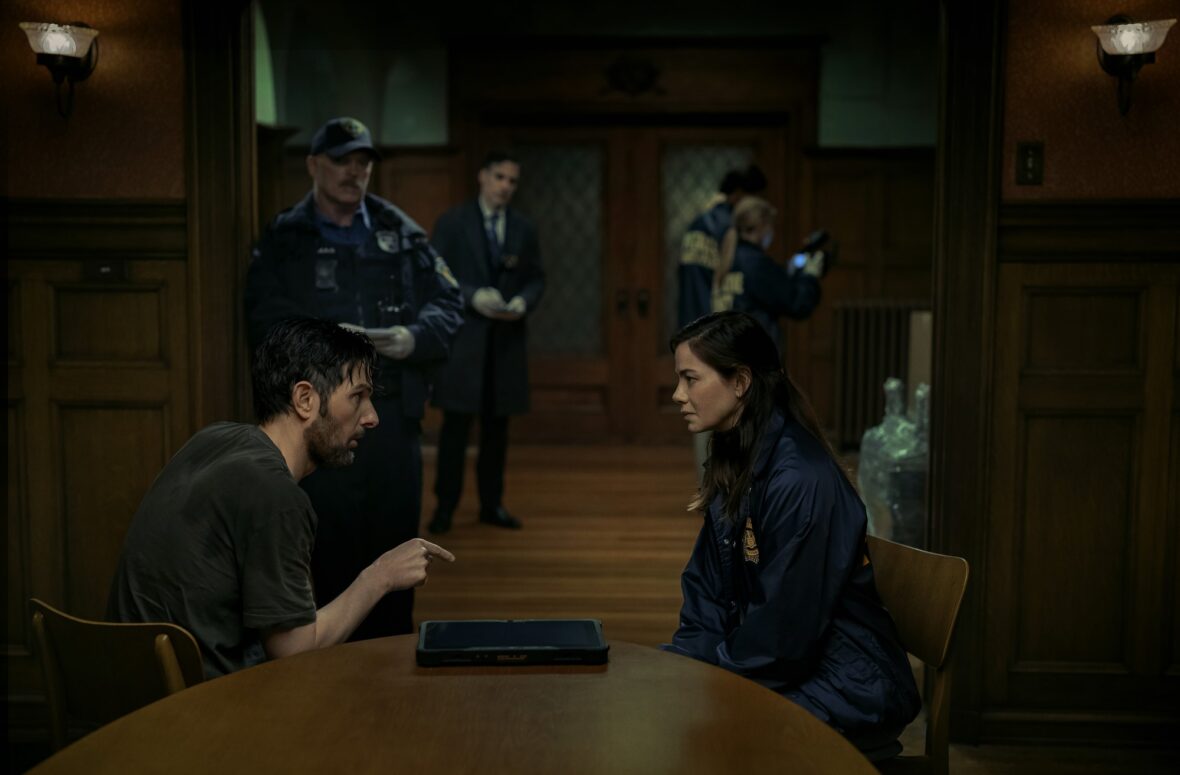Adam Scott and Michelle Monaghan sit across from a table with one another as police appear in the background in still from