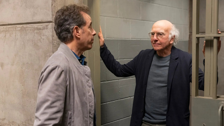 Larry (Larry David) and Jerry (Jerry Seinfeld) stand in a gray jail cell doorway with Larry wearing glasses and gripping the bars while Jerry faces him in profile both with serious expressions in Curb Your Enthusiasm